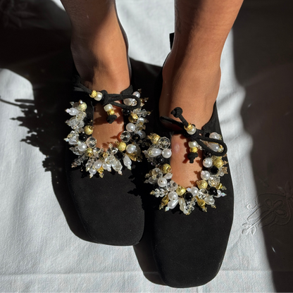 Black flat shoes with ornate crystal and pearl embellishments and bow details, shown on feet against a white fabric background.